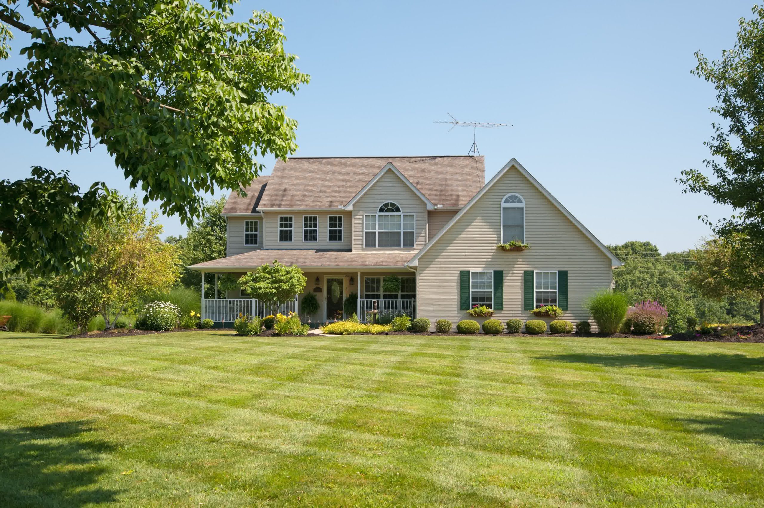 Suburban two-story beige house with green shutters, landscaped yard, striped lawn, and large front porch.