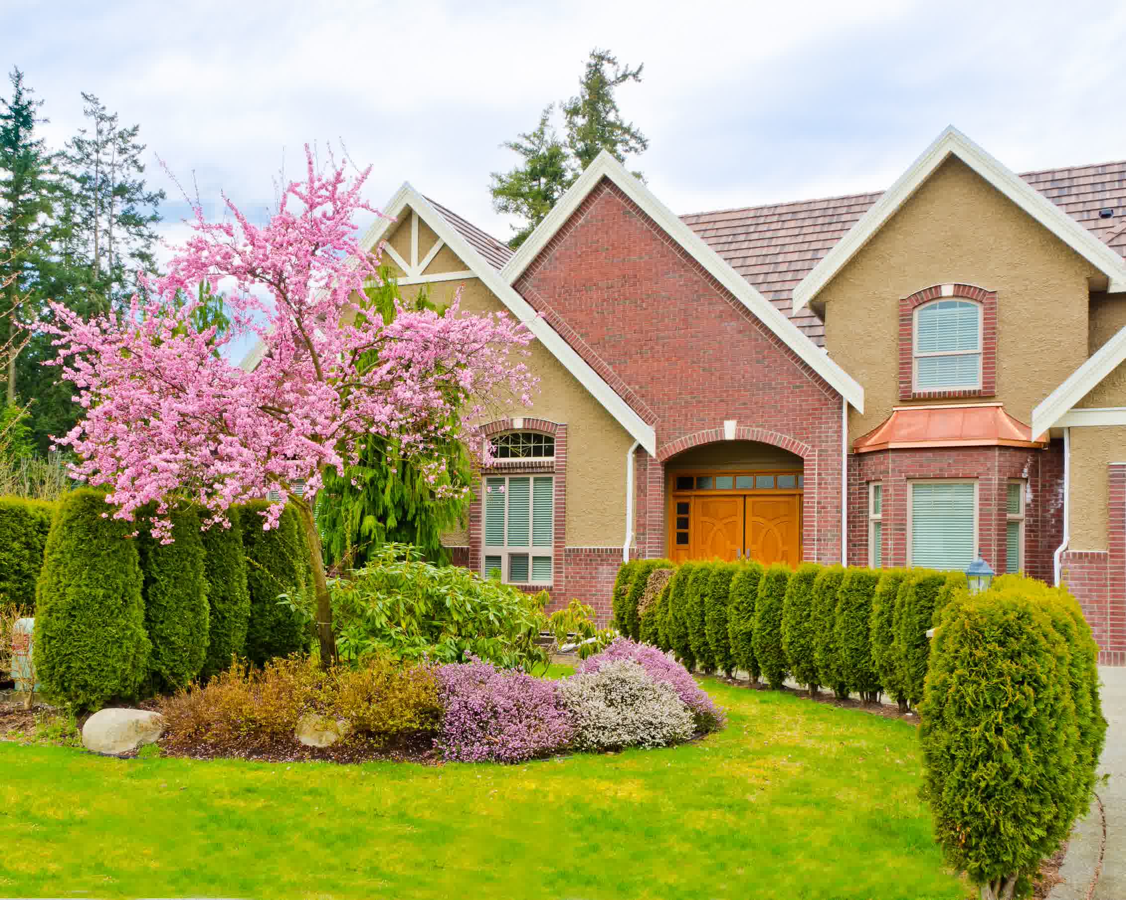 Brick home with blooming pink cherry blossom tree, trimmed hedges, and manicured lawn in a colorful spring landscape design.