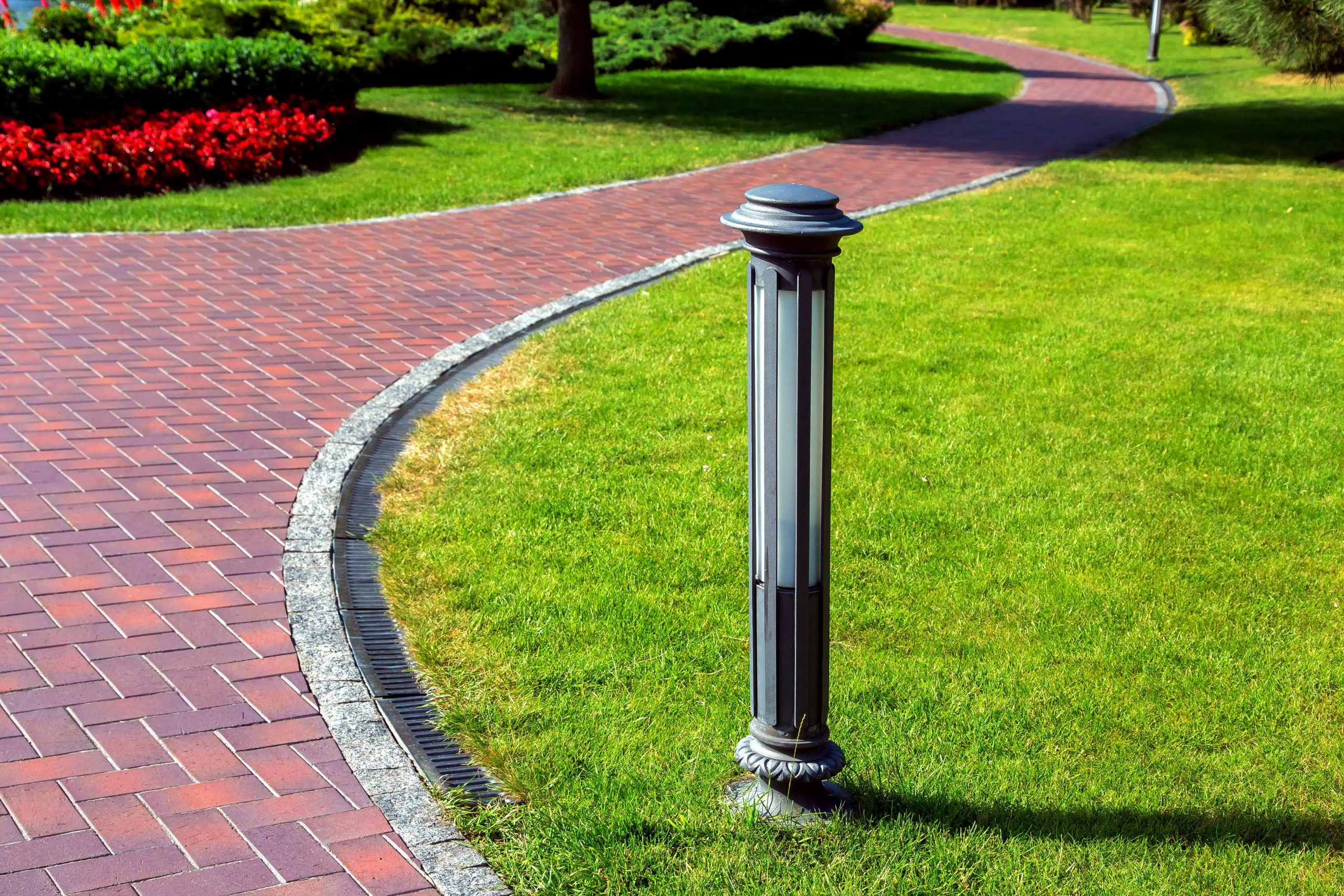 Curved red brick walkway with decorative edging and garden lighting winds through green grass and flower beds in a landscaped park.