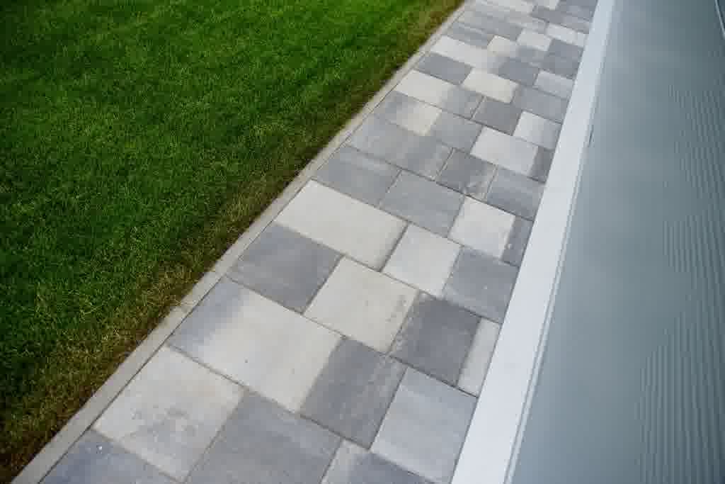 Modern stone paver walkway with gray and white tiles runs beside a green lawn and home exterior, creating a clean landscape design.