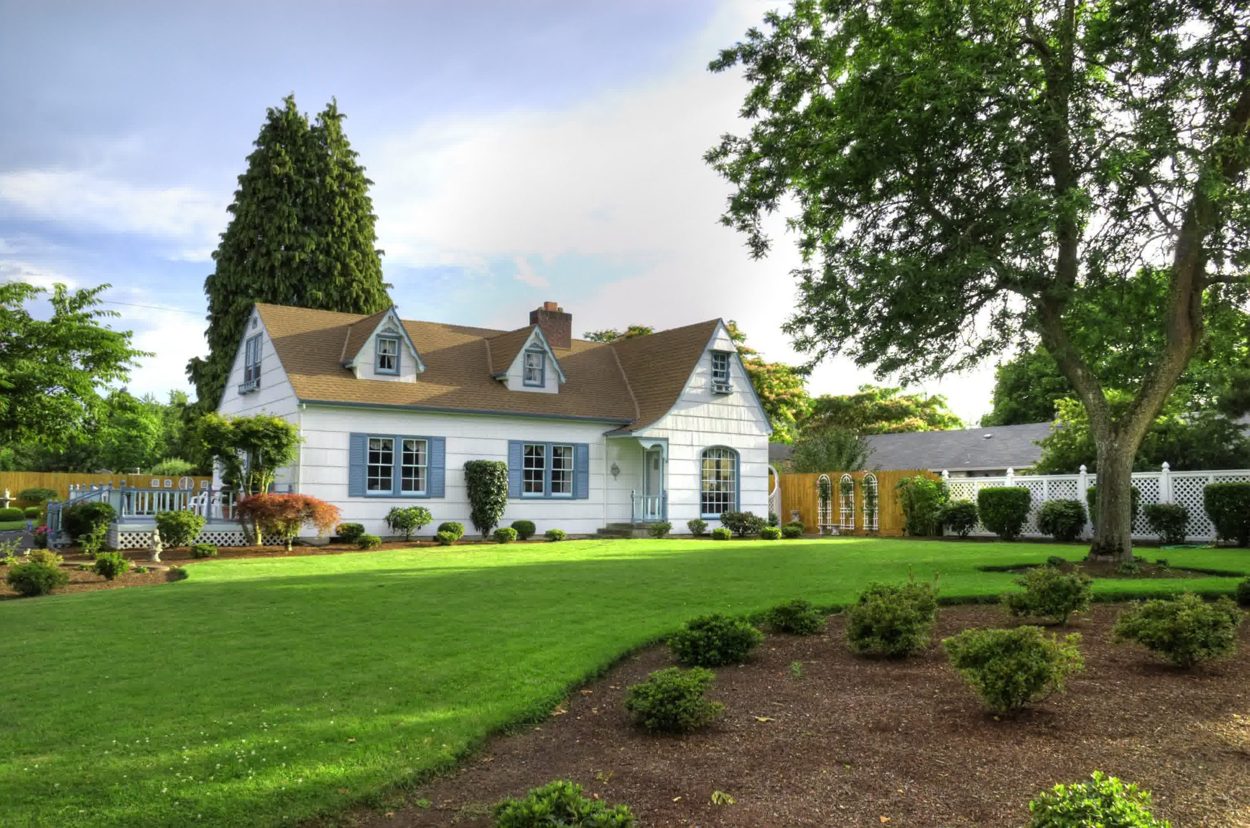 Charming white cottage-style home with blue shutters, manicured lawn, garden beds, and mature trees in a landscaped suburban yard.