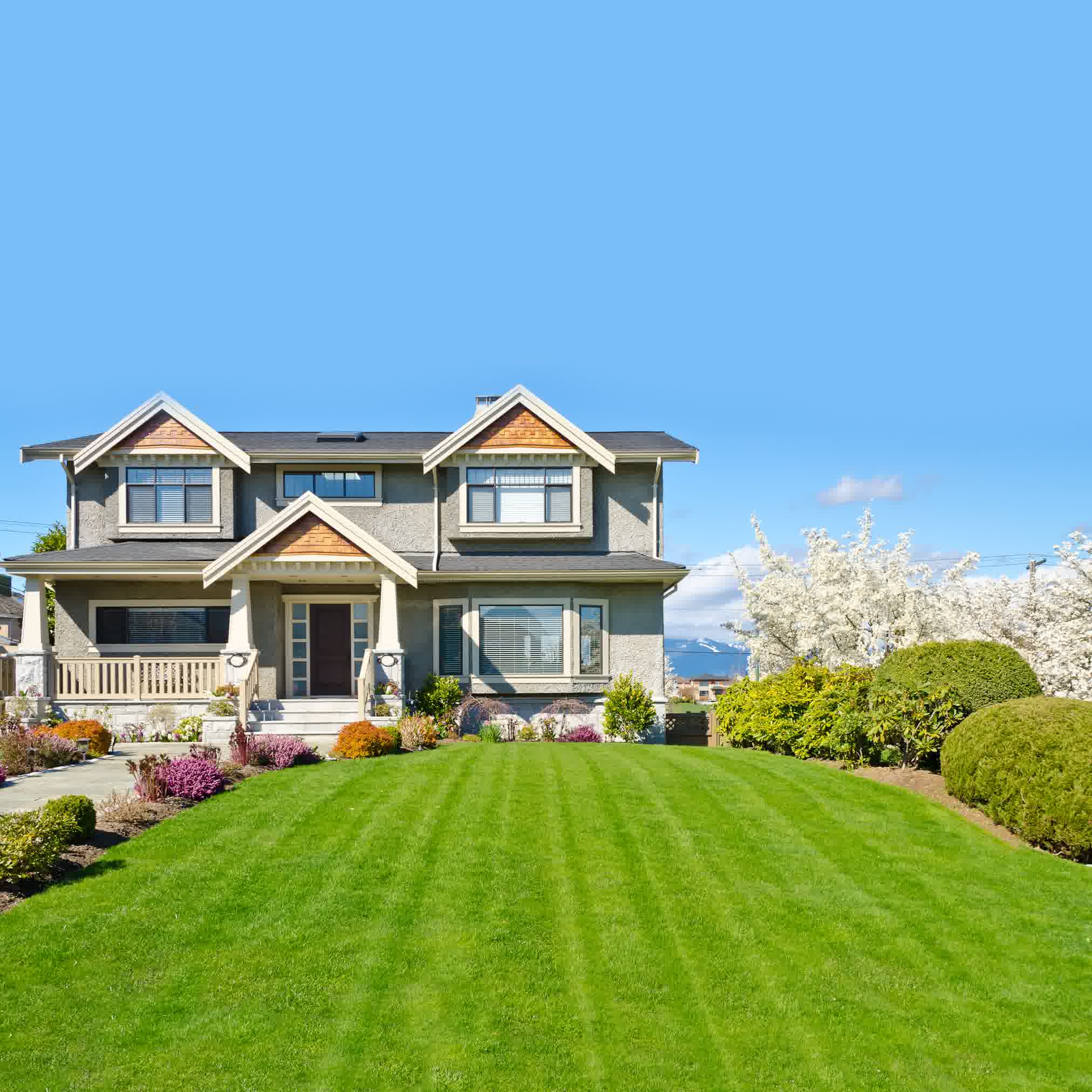 Modern gray two-story home with gabled roof, lush green lawn, colorful flower beds, and blooming white tree.
