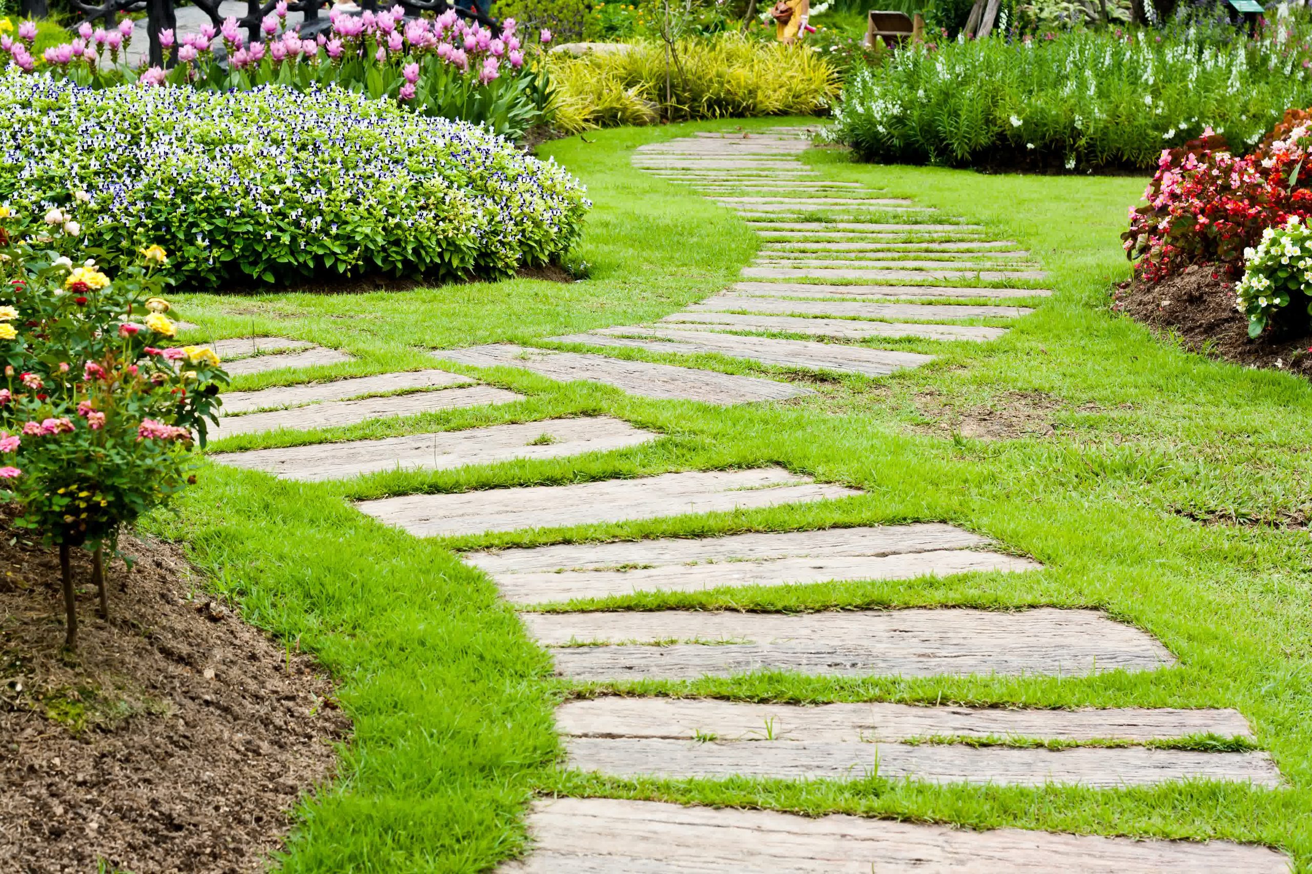 Curved stone garden pathway surrounded by vibrant flower beds with pink, purple, and yellow blooms in a landscaped garden.