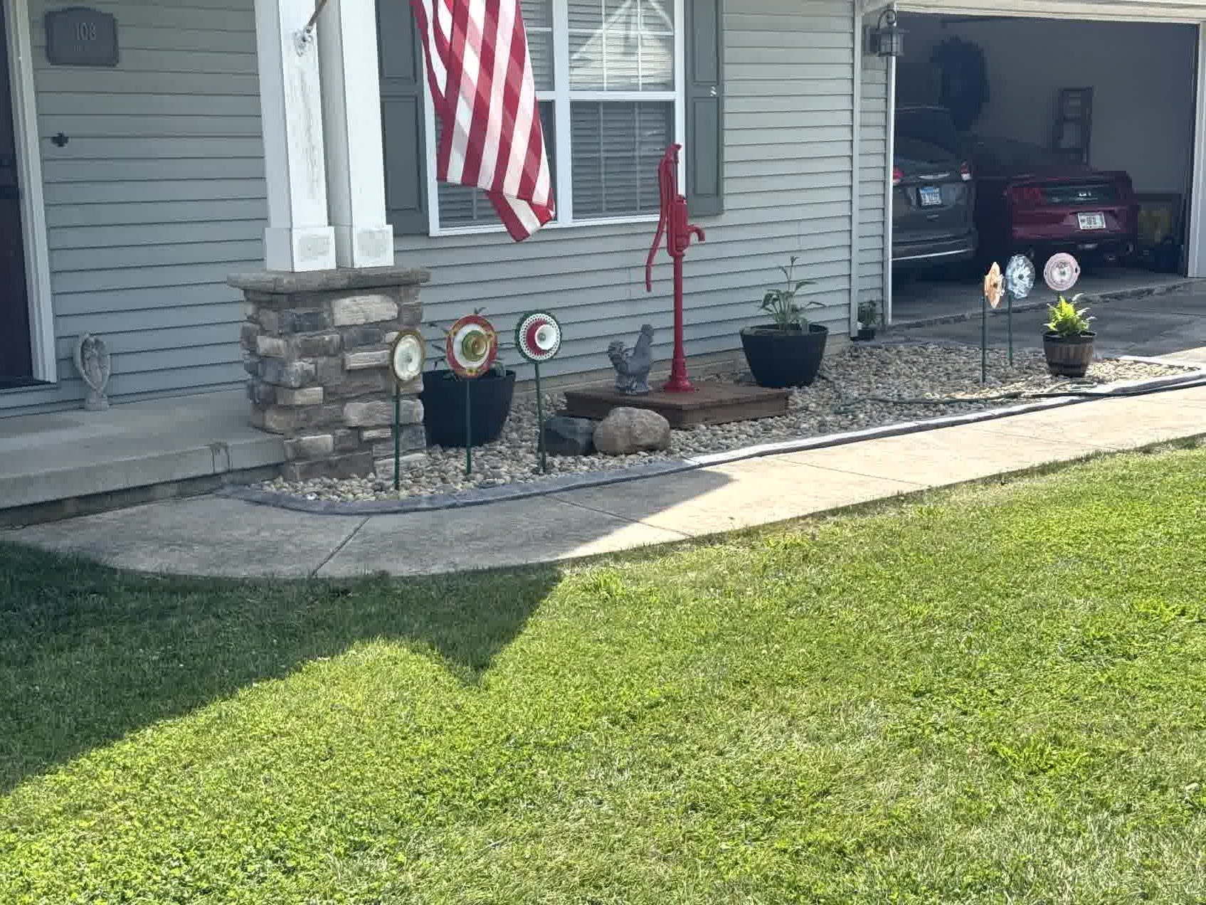 Front porch landscape with stone edging, red water pump decor, flower pots, and plate art in rock bed near concrete walkway.
