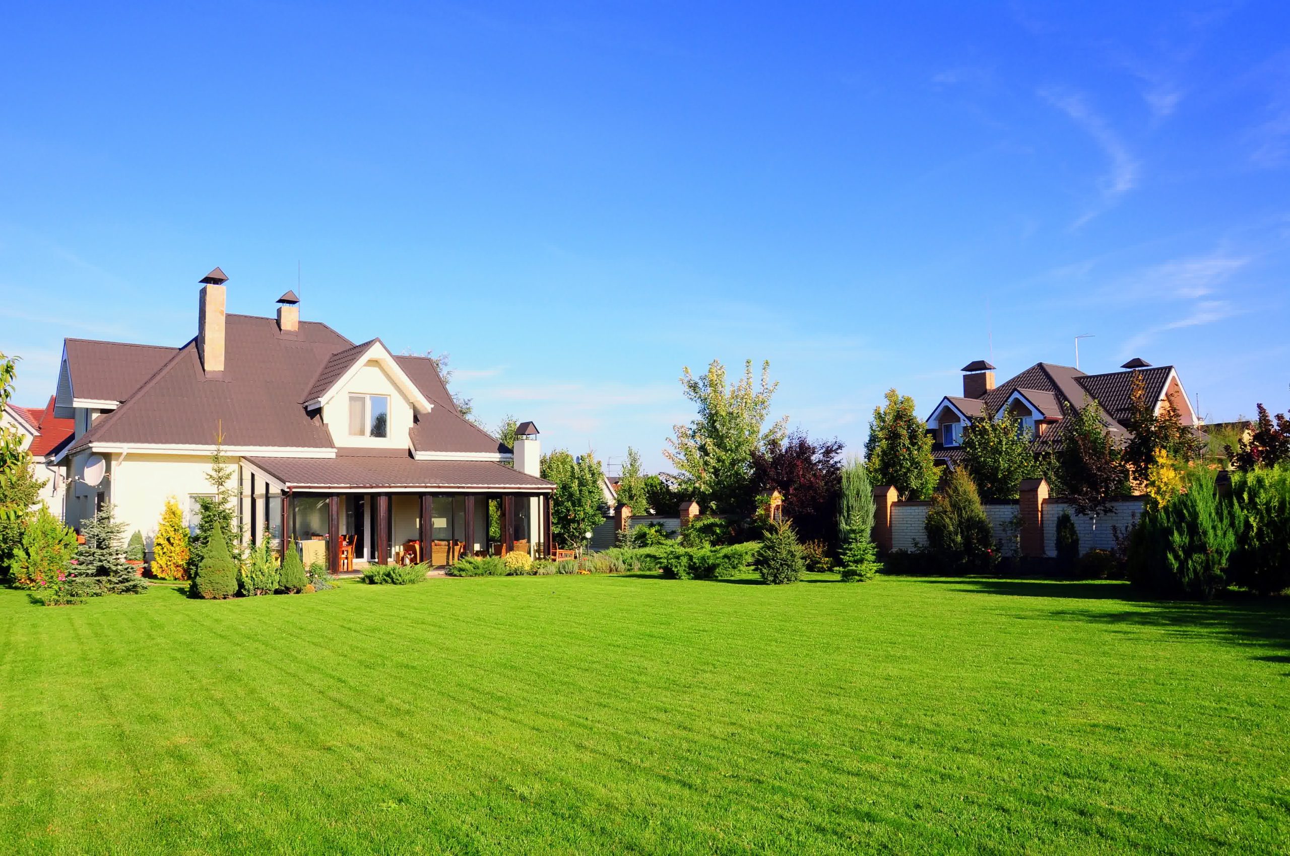 Large country house with brown roof, wide lawn, manicured garden, and patio seating under clear blue sky.
