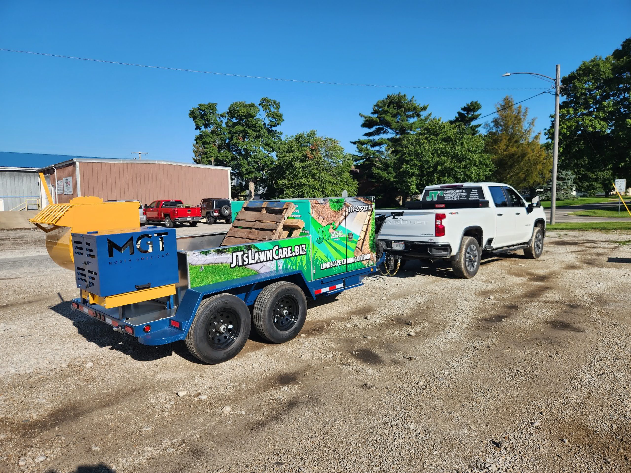 White pickup truck towing a landscaping trailer with lawn care equipment and branding parked on a gravel lot near industrial buildings.
