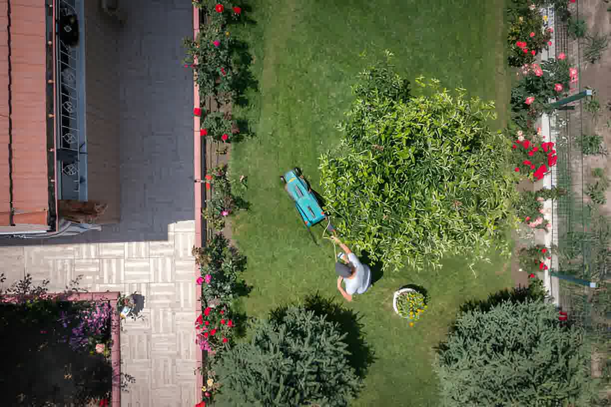 Aerial view of a person mowing a lush green backyard lawn bordered by flower beds and ornamental shrubs near a tiled patio.